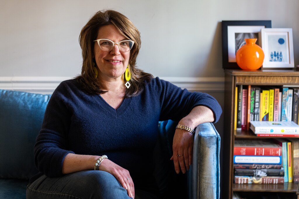 Stephanie Mellinger smiles for a portrait while sitting on a couch near a bookshelf.