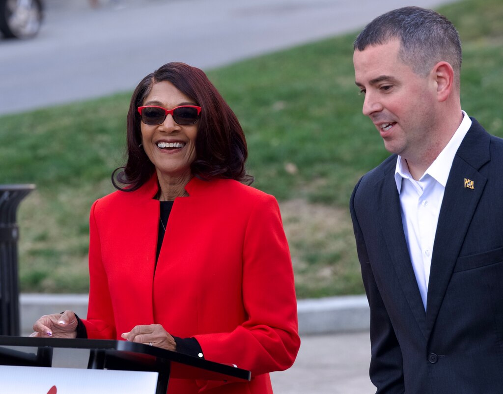 Sheila Dixon stands behind a lectern in a red suit jacket, wearing sunglasses and smiling broadly. To her left, Eric Costello wears a dark suit and white shirt.