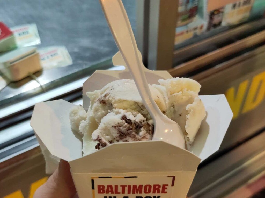 A small white box that says "BALTIMORE IN A BOX" on the front holds ice cream and a spoon in front of an open window.