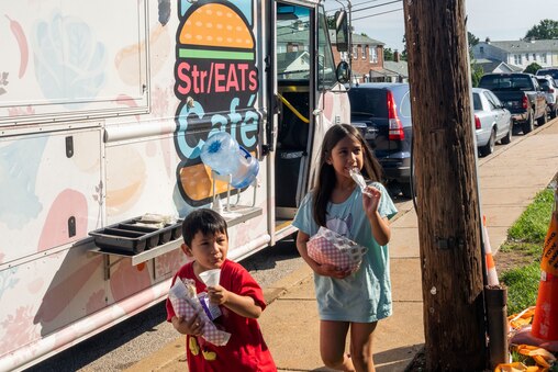 Julieta Salazar-Bueso and little brother José are accompanied by their mother Ruth and baby sibling as they recieve breakfast from the Streats Cafe food truck outside of Armistead Gardens Elementary/Middle School on July 3, 2025.