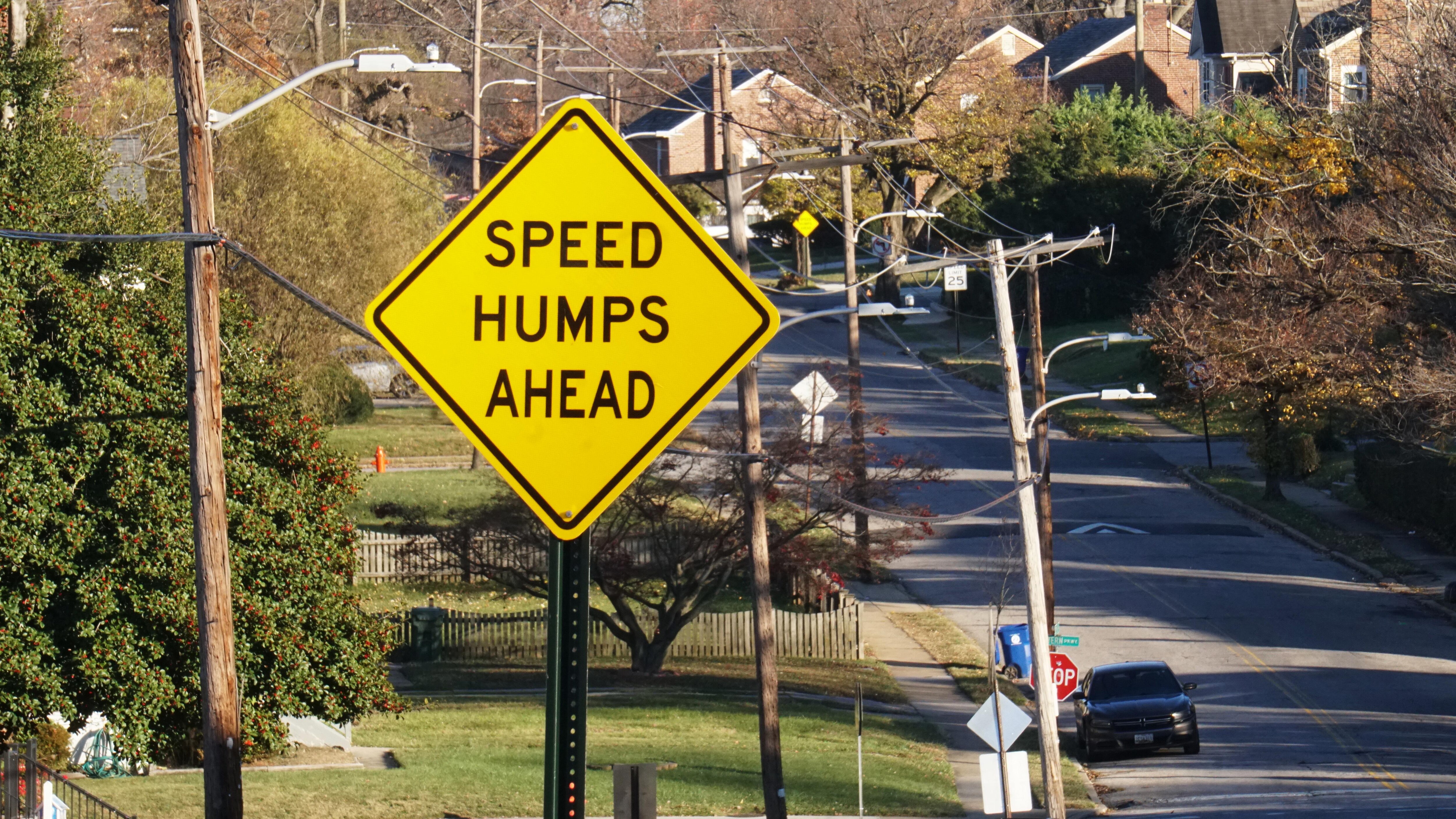 A yellow street sign that says "SPEED HUMPS AHEAD" in front of a roadway with a speed hump in the background.