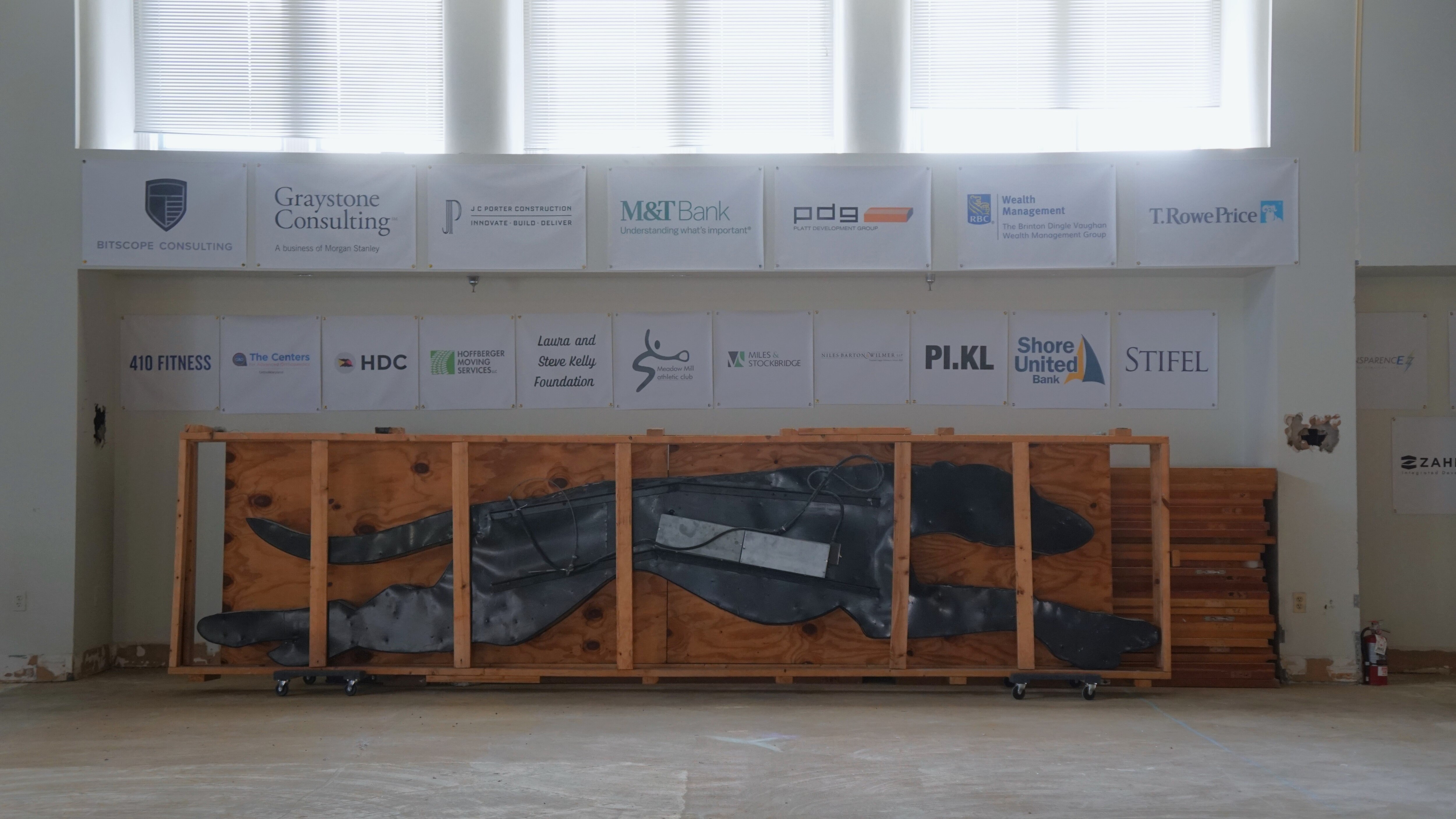 An old Greyhound bus logo sign is boarded up in a wooden transport frame. It rests inside an office-like building underneath a long window.