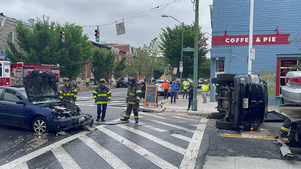 Fire fighters standing in an intersection next to a blue car involved in a crash. An overturned car is on the other side of the crosswalk.