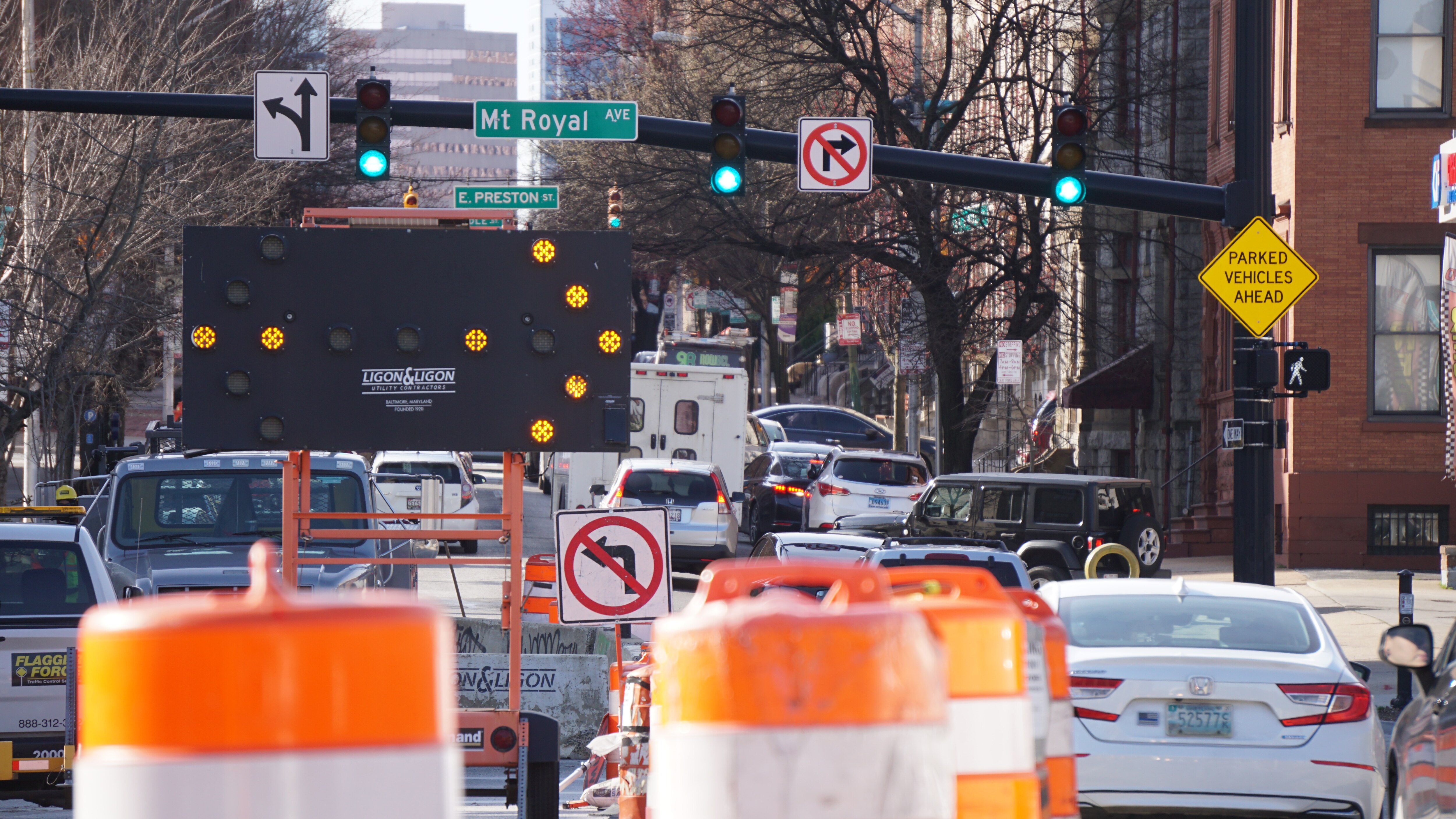 A mobile roadway sign illuminates a directional arrow to indicate where a line of cars should travel. Orange roadwork barriers are in the foreground, a street sign that says 'Mt. Royal Ave' hangs next to a green traffic light above the road.