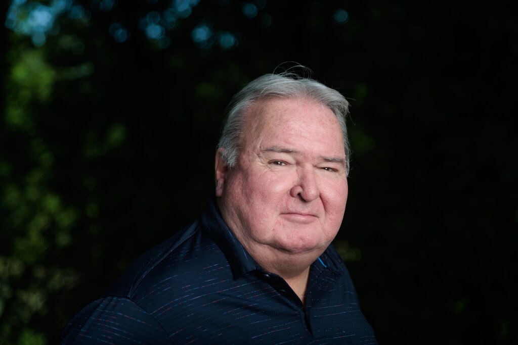 Photograph of older white man with silver hair and wearing a dark collared shirt, looking into the camera. The background is dark with some blurry patches of sky through the tree canopy.