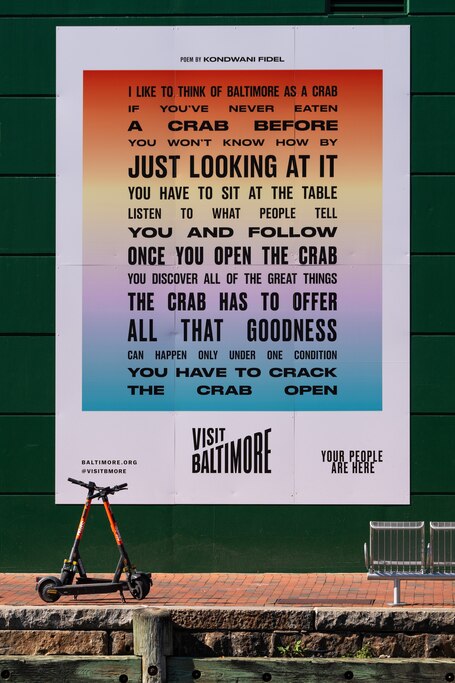 Two orange Spin scooters are parked in front of a colorful, linear gradient Visit Baltimore poster that has a poem by Kondwani Fidel on it.