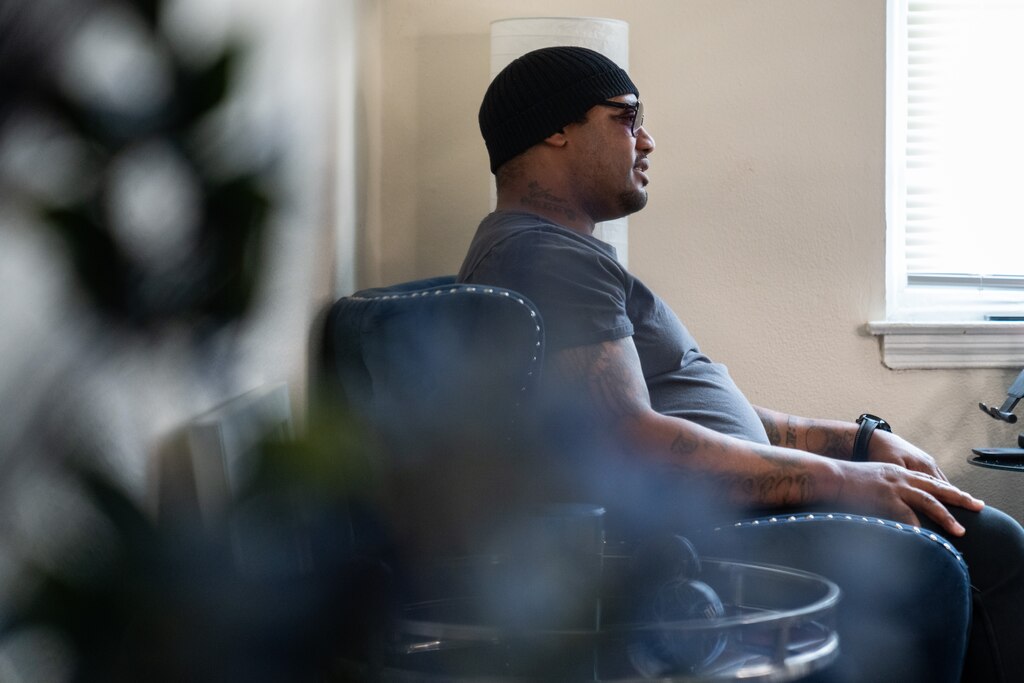 A side profile of Nathaniel Powell, wearing sunglasses, as he speaks while sitting in a blue velvet chair in his home.