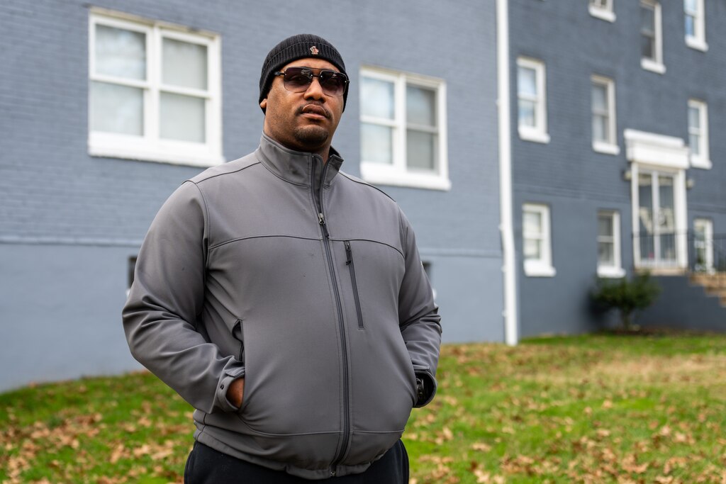 Nathaniel Powell, wearing sunglasses, stands for a portrait outside of a blue apartment building with his hands in his pockets.