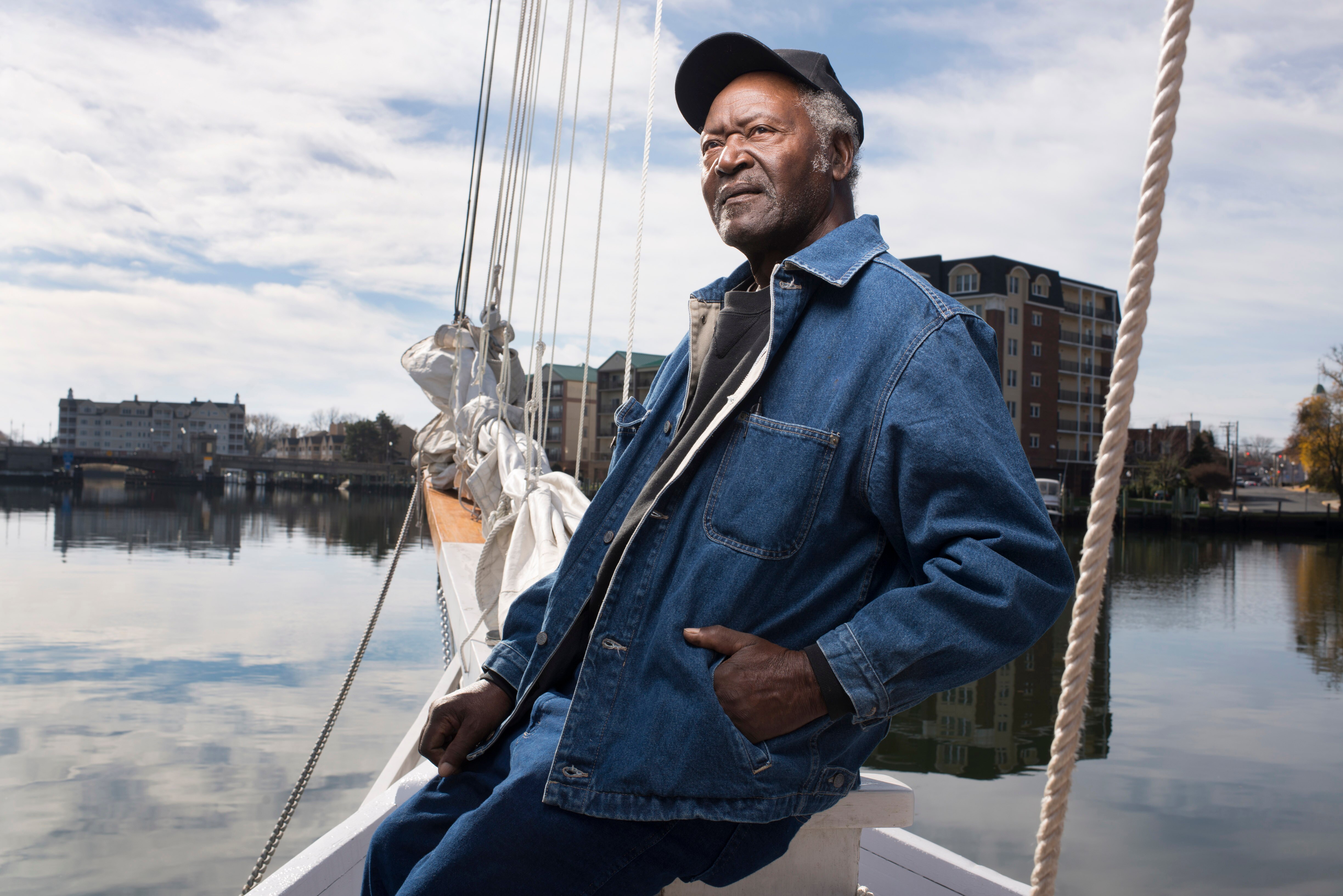 Kermit Travers poses on the stern of a sailboat with water and buildings in the background. He wears a black baseball cap, a blue denim jacket and a black shirt as he looks up and away from the lens.