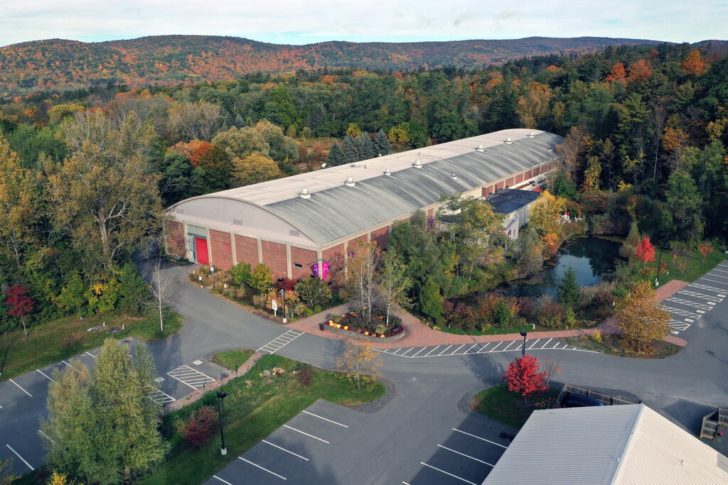 Aerial photo of long rectangular building with arched roof nestled among autumnal trees and hills in the background, and parking lot in front.