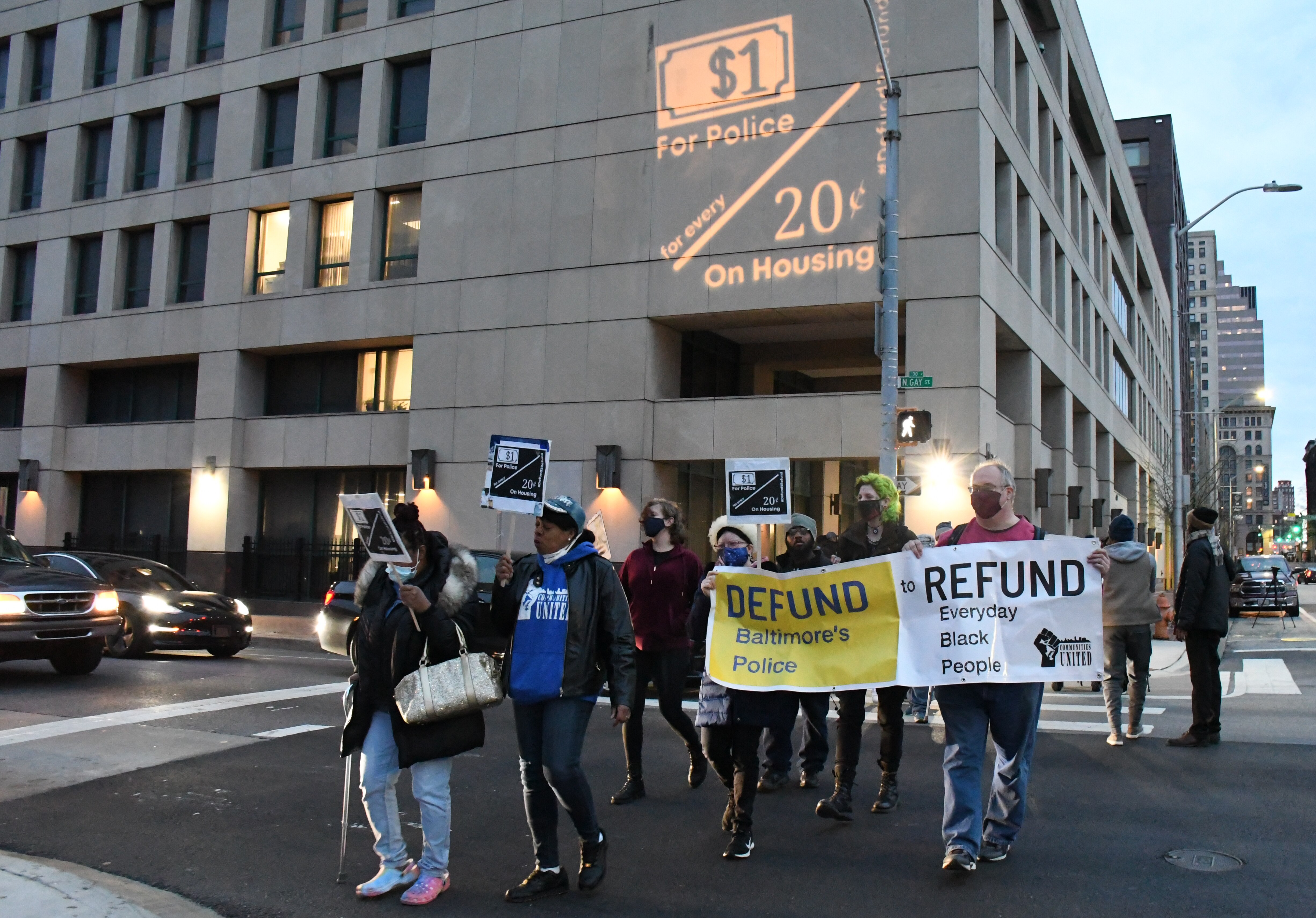 Photo by Pamela Wood/The Baltimore Banner -- Demonstrators organized by Communities United march briefly outside the Charles R. Benton Jr. Building in Baltimore on Wednesday, March 30, 2022 to call attention to disparities in government spending on police versus housing. They used a projector to put their message on the side of the building, where the offices of the city's housing authority are located.
