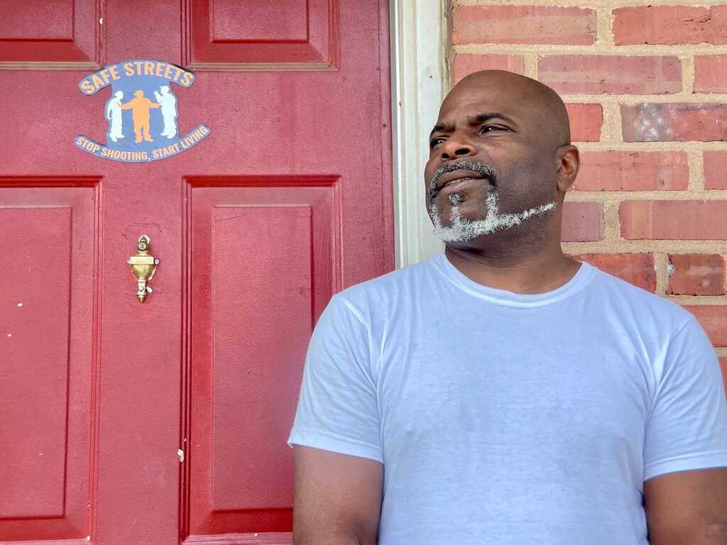 A man in a T-shirt with a white beard stands in front of a red door with a Safe Streets sticker on it.