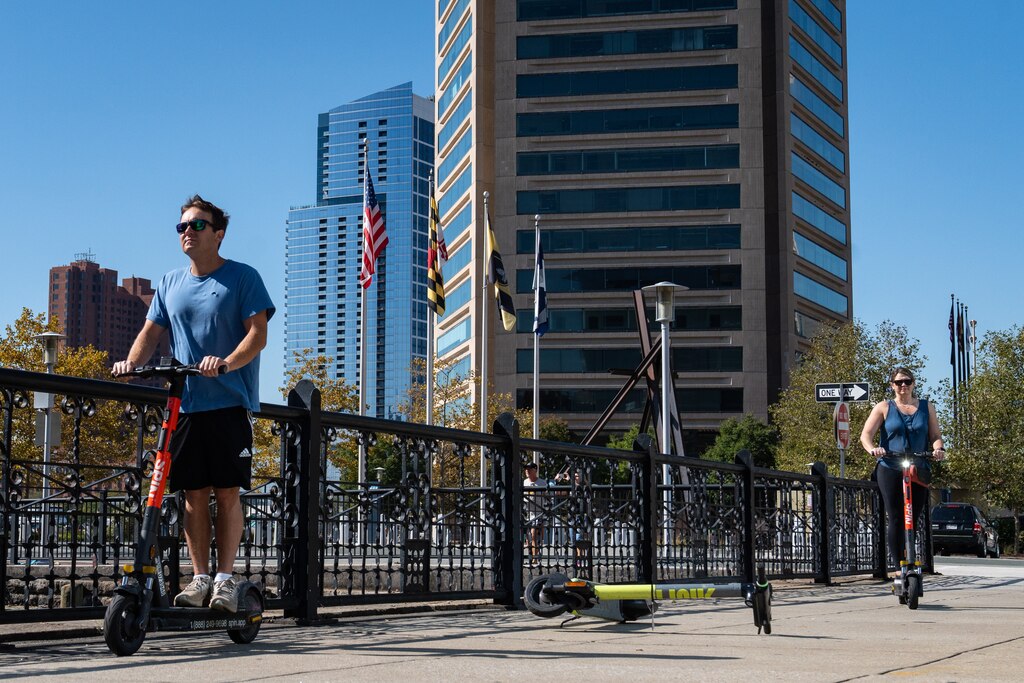 A man and woman scooter through Inner Harbor. The Baltimore World Trade Center is behind them and an unused scooter lies on the ground between them.