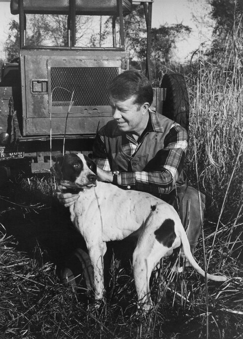 American politician Jimmy Carter crouching down to pet his dog behind an tractor on his peanut farm in Plains, Georgia, 1970. Carter served two terms as a Georgia State Senator from 1963 to 1967, and one as the Governor of Georgia from 1971 to 1975, prior to his election as the 39th President of the United States from 1977 to 1981.