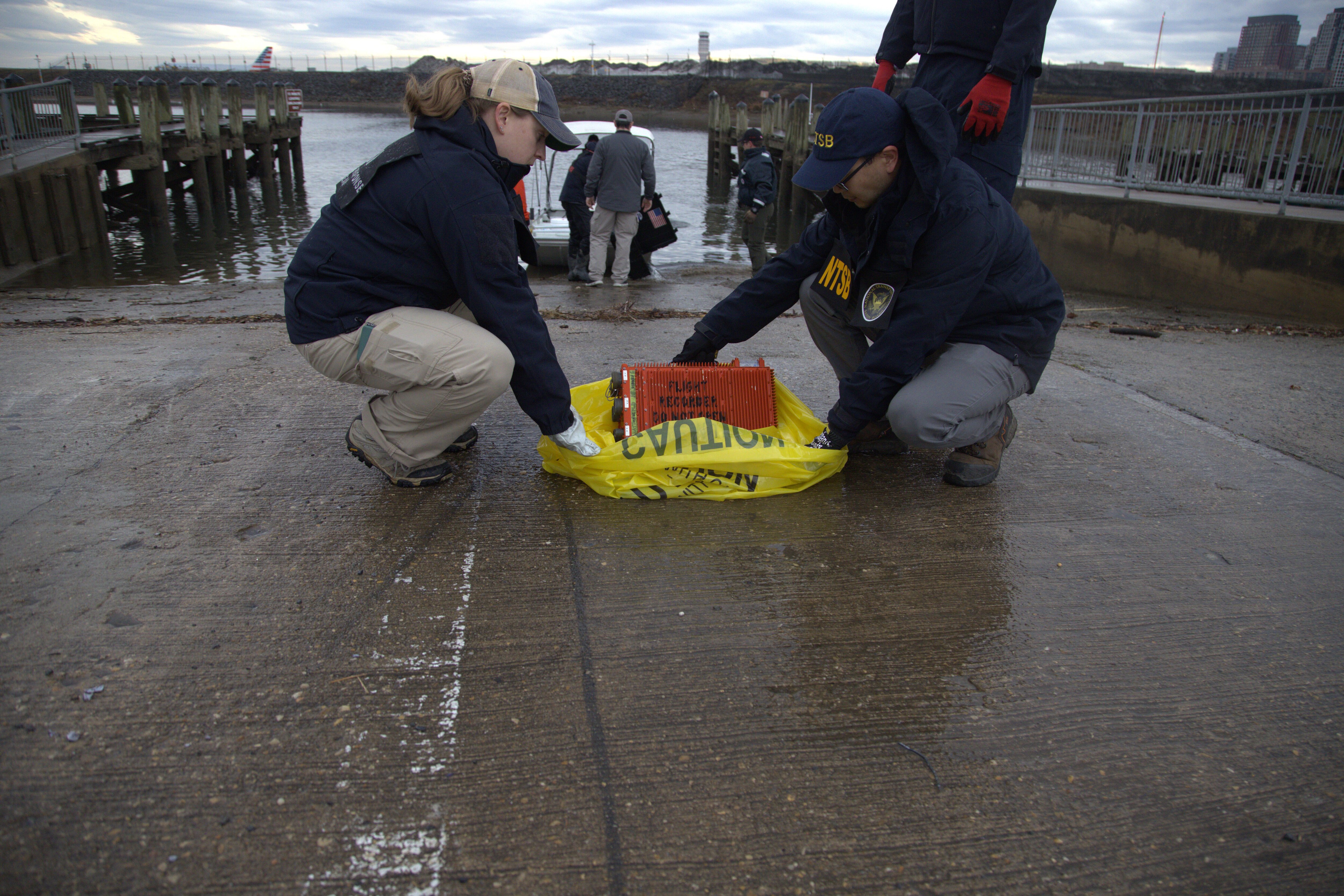 An orange flight data and cockpit voice recorder box sits on a yellow bag labeled "CAUTION." Two NTSB investigators are holding the edges of the bag and starting to lift it over the box.