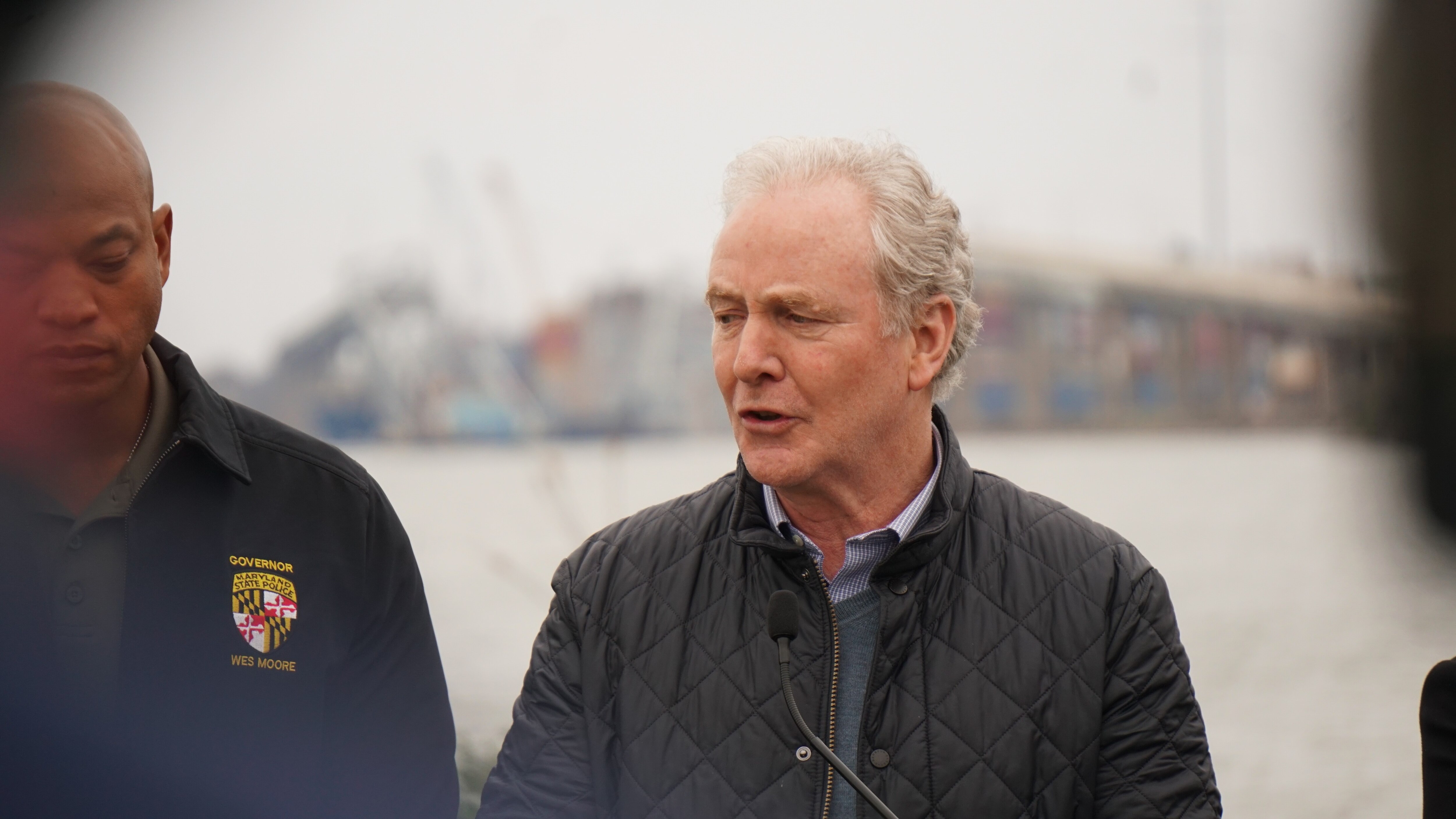 A man speaks into a podium microphone outside and the remains of a bridge are seen in the background.