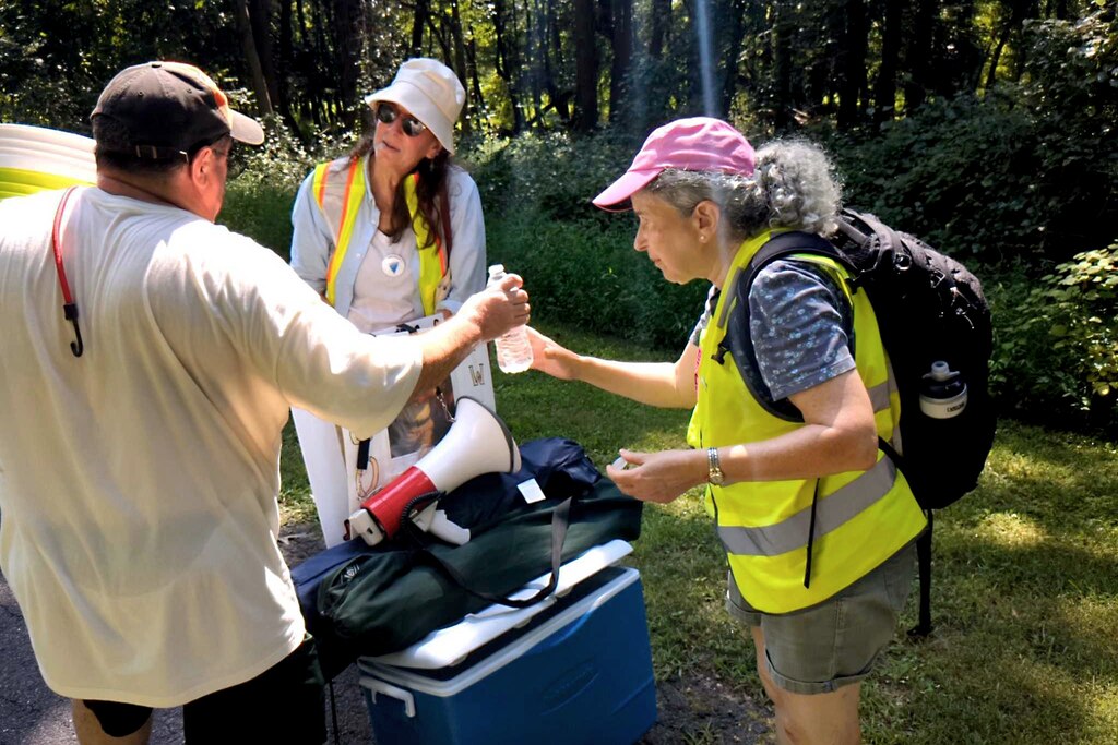 People gathered Sunday, July 27, 2025, on an Interstate 195 overpass near BWI Thurgood Marshall Airport to protest Avelo Airlines deal with ICE to operate deportation flights. Avelo operates a limited number of passenger flights out of BWI. Dr. Kate Sugarman, right, hands a bottle of water to a protester. The heat index topped 100 degrees.