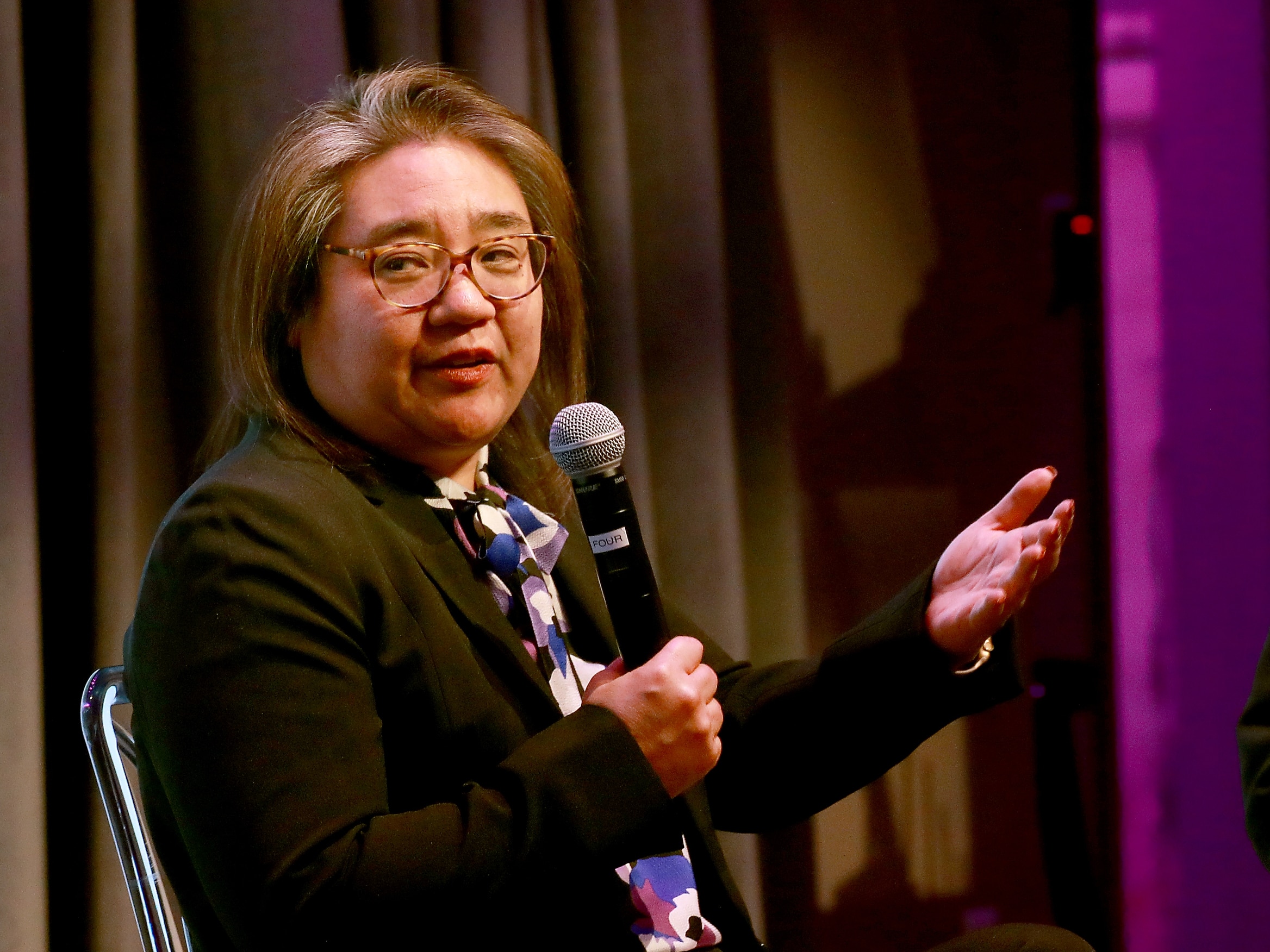 Kimi Yoshino holds a microphone in her hand as she speaks to an audience. She is the only person in the photo and is sitting in a metal chair in front of a black curtain.