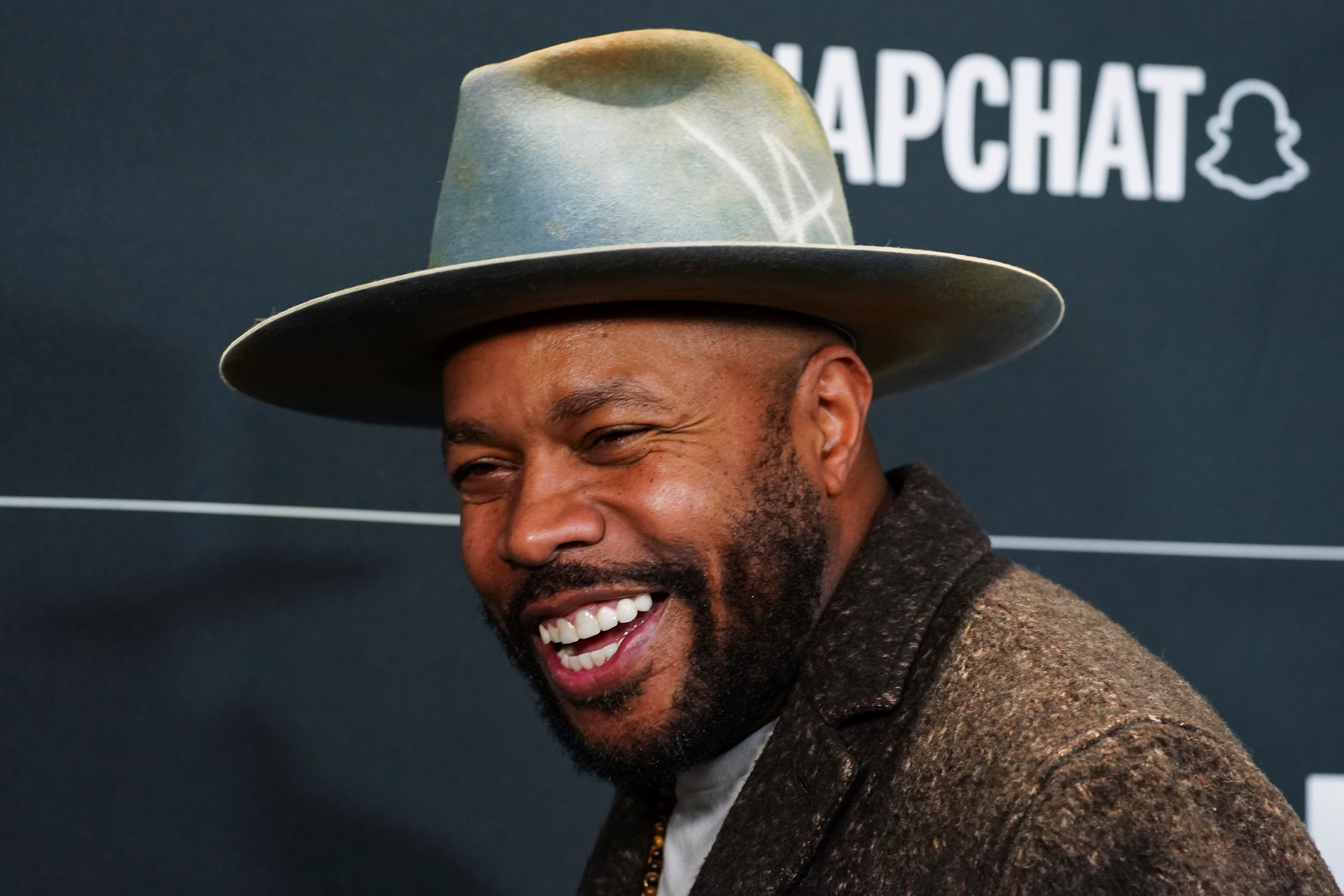 D-Nice appears in a wide-brimmed hat and smiling in front of a dark backdrop.